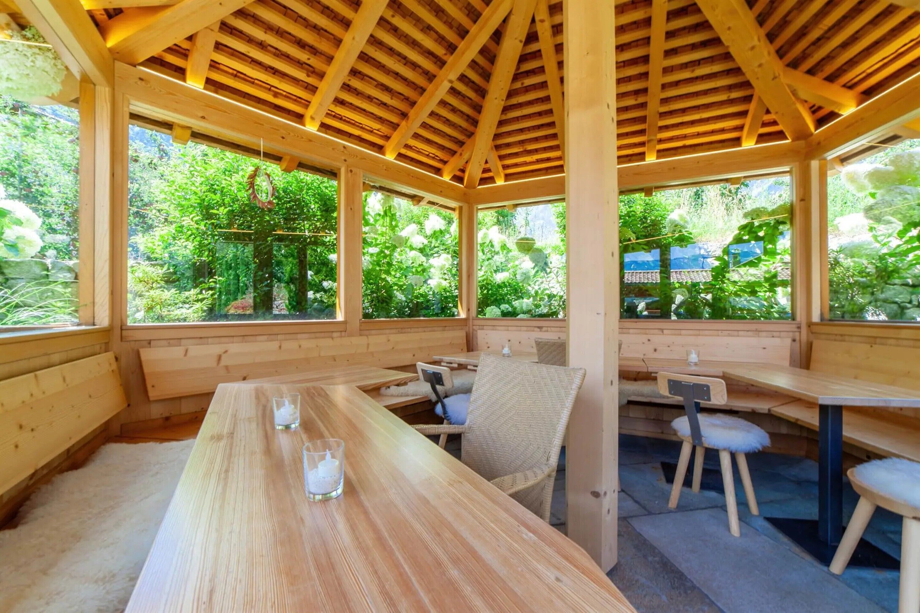 Ein hölzernes Pavillon mit Bänken und Tischen, großen Fenstern und Blick ins Grüne.