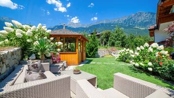 Eine Terrasse mit Korbmöbeln, einem hölzernen Pavillon und einem gepflegten Rasen mit Blumen.
