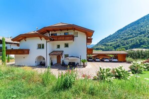 A two-story building with a balcony, a covered outdoor seating area, and a driveway with cars. - Apartment “Tratter 4°” with Mountain View, Shared Garden and Wi-Fi (Lagundo)