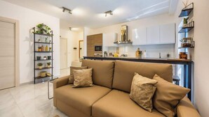 A modern living room with a beige sofa, a kitchen with white cabinets, and a small shelf with plants. - Palazzo Ducoss - Apt 7 - Two Bedroom One Bathroom (Valletta)