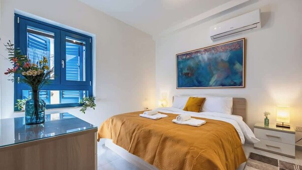 A bedroom with a bed, bedside tables, a nightstand, a window with blue shutters, an air conditioner, and a painting on the wall. - Palazzo Ducoss - Apt 7 - Two Bedroom One Bathroom (Valletta)