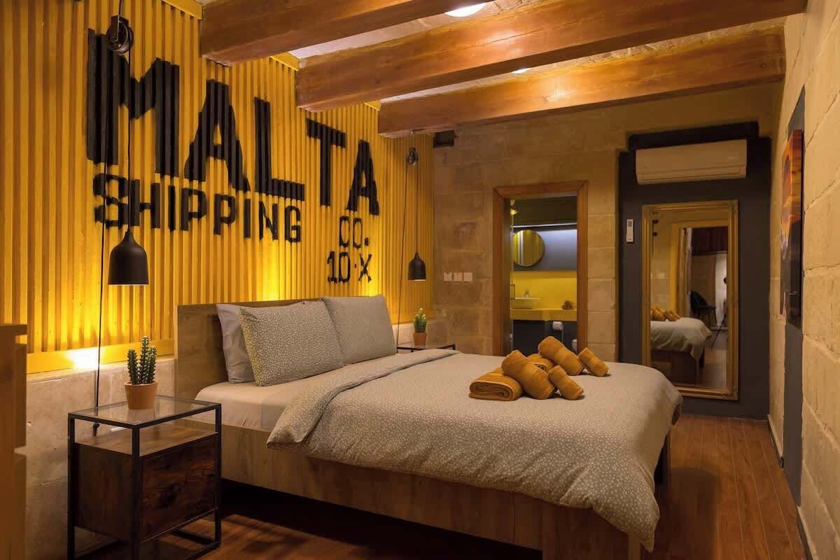 A bedroom with a bed, a bedside table, a potted plant, and a wall with 'MALTA SHIPPING' text.