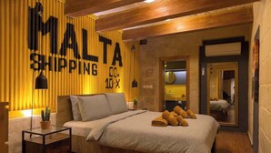 Makuuhuone, jossa on sänky, yöpöytä, ruukkukasvi ja seinä, jossa on teksti ”MALTA SHIPPING”.