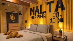 Makuuhuone, jossa on sänky, tyynyjä, yöpöytä, lamppu ja seinä, jossa lukee teksti: ”MALT SHIPPING”.