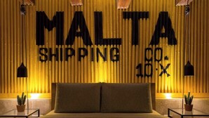 Huoneessa on sänky, seinässä teksti ”MALTA SHIPPING CO. LTD” ja kaksi ruukussa olevaa kasvia.