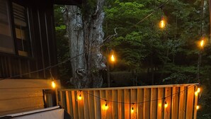 Un jacuzzi en una terraza de madera con luces colgantes, rodeado por una valla de madera y árboles.