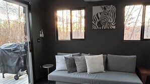Una sala moderna con un sofá gris, un otoman redonda y un tapete estampado. Varias ventanas ofrecen luz natural, y un cuadro con estampado de cebra adorna la pared.