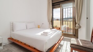 Ett hotellrum med en säng, en stol, ett litet bord och en balkong med gardiner.