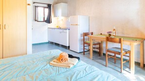 A bedroom with a bed, a wooden wardrobe, a kitchenette, and a dining area with a table and chairs. - Studio Apartment 'Rico Plage 2 Personnes' with Private Terrace, Wi-Fi and Air Conditioning (Talasani)