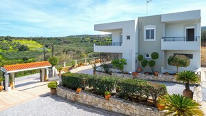 A modern two-story building with a stone retaining wall, potted plants, and a clear view of the surrounding landscape. - Villa Pantelis with private pool (Rethymno)