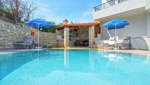 A swimming pool with lounge chairs and umbrellas, a stone structure with a chimney, and a modern building in the background. - Villa Pantelis with private pool (Rethymno)