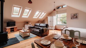 A cozy loft bedroom with a large bed, a leather sofa, a dining table set for two, and a TV in a corner. - Ellenboro House, Cosy Sleeps 2, Near Cartmel (Newton-in-Cartmel)