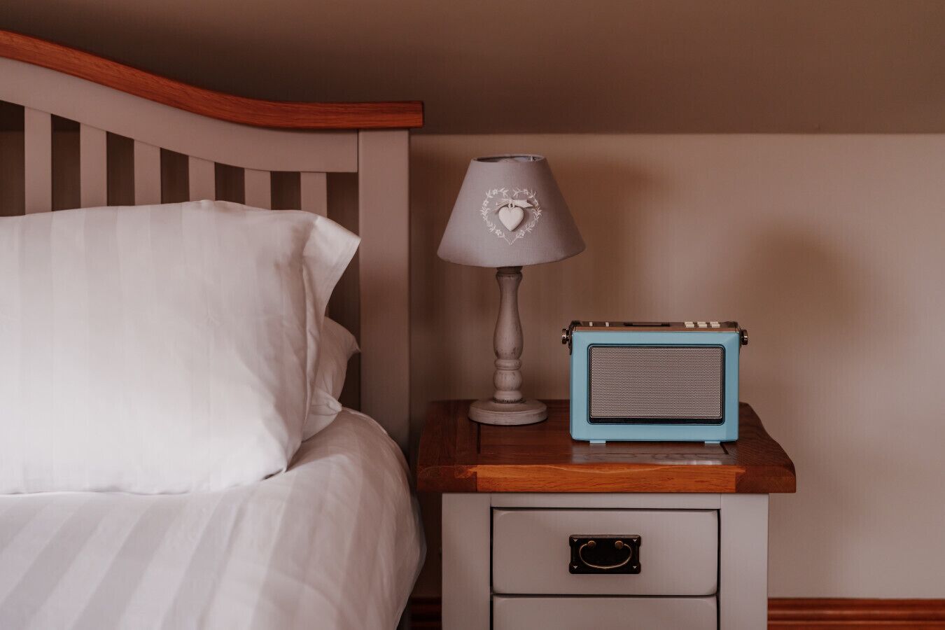 Ein Bett mit weißer Bettwäsche, eine Nachttischlampe mit Herzmotiv und ein blaues, altes Radio auf einem Nachttisch aus Holz mit einer Schublade.