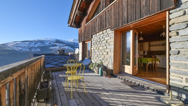 Eine Holzterrasse mit Tisch und Stühlen, eine Steinmauer und Blick auf einen verschneiten Berg.