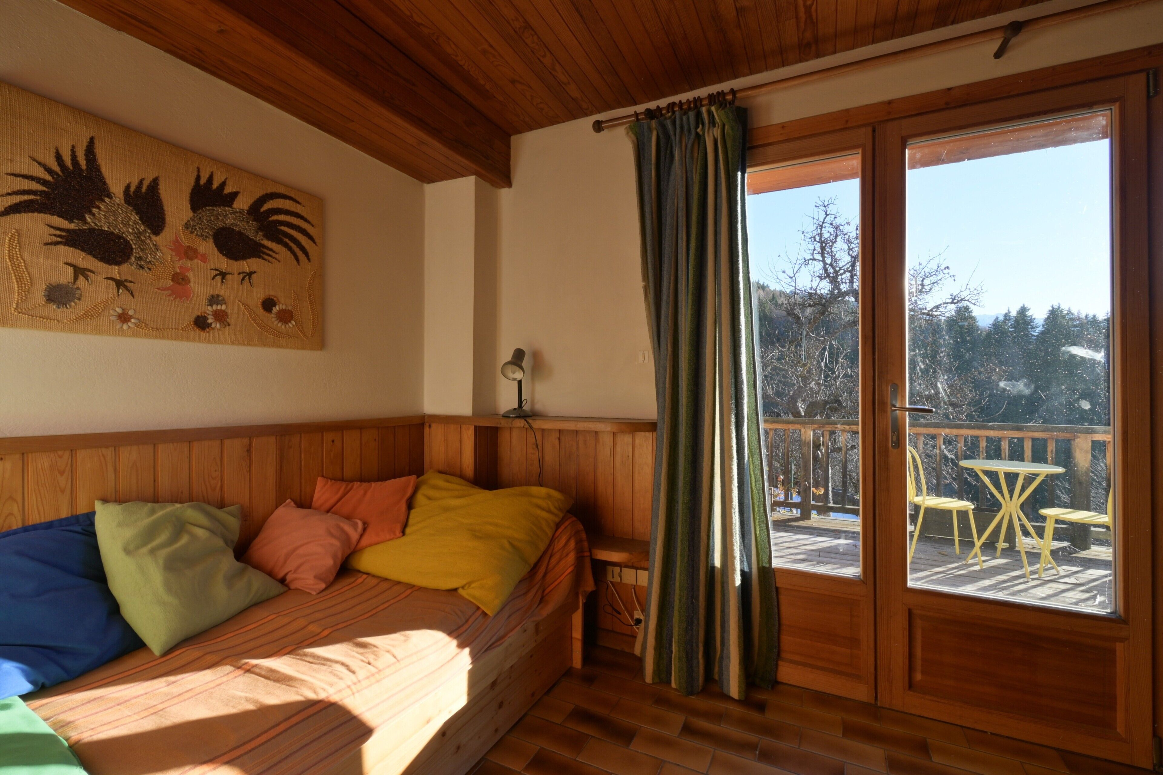 Une pièce de cabane en bois comprenant un lit, une lampe fixée au mur, une fenêtre avec des rideaux et une vue sur une terrasse avec des chaises.