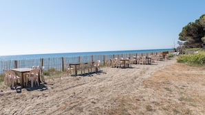 A beachside setting with tables and chairs arranged for dining, a wooden fence, and a clear view of the sea. - Apartment 'Rico Plage 4 Personnes' with Private Terrace, Wi-Fi and Air Conditioning (Talasani)