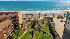 A coastal hotel with a view of the ocean, a parking lot, and a small garden area with palm trees. - Seashell Retreat- Beachfront condo/pools/cabanas (Galveston)