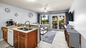 A compact kitchen with a sink, stove, and cabinets, a dining area with a table and chairs, and a balcony with a view of palm trees. - Seashell Retreat- Beachfront condo/pools/cabanas (Galveston)