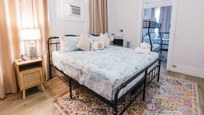 A bedroom with a bed, bedside table, lamp, air conditioning unit, and a view of a balcony.