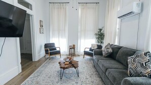 A living room with a grey sofa, a wooden coffee table, and two armchairs. There is a flat-screen TV mounted on the wall, a potted plant, and a window with sheer curtains.