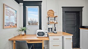 Een kleine keuken met een houten tafel, witte kastjes, een zwarte deur en een raam met uitzicht op bomen.
