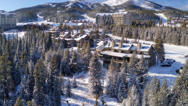 Estância coberta de neve com vários edifícios, incluindo um grande chalé e algumas cabanas menores, rodeada por florestas densas e uma montanha ao fundo.