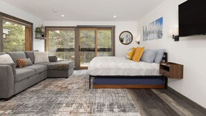 Quarto moderno com cama grande, sofá seccional cinza e vista através de uma porta de vidro deslizante.