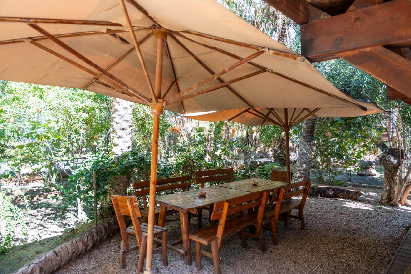 An outdoor wooden table and chairs under a large umbrella, surrounded by greenery.