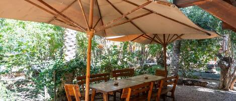 An outdoor wooden table and chairs under a large umbrella, surrounded by greenery.