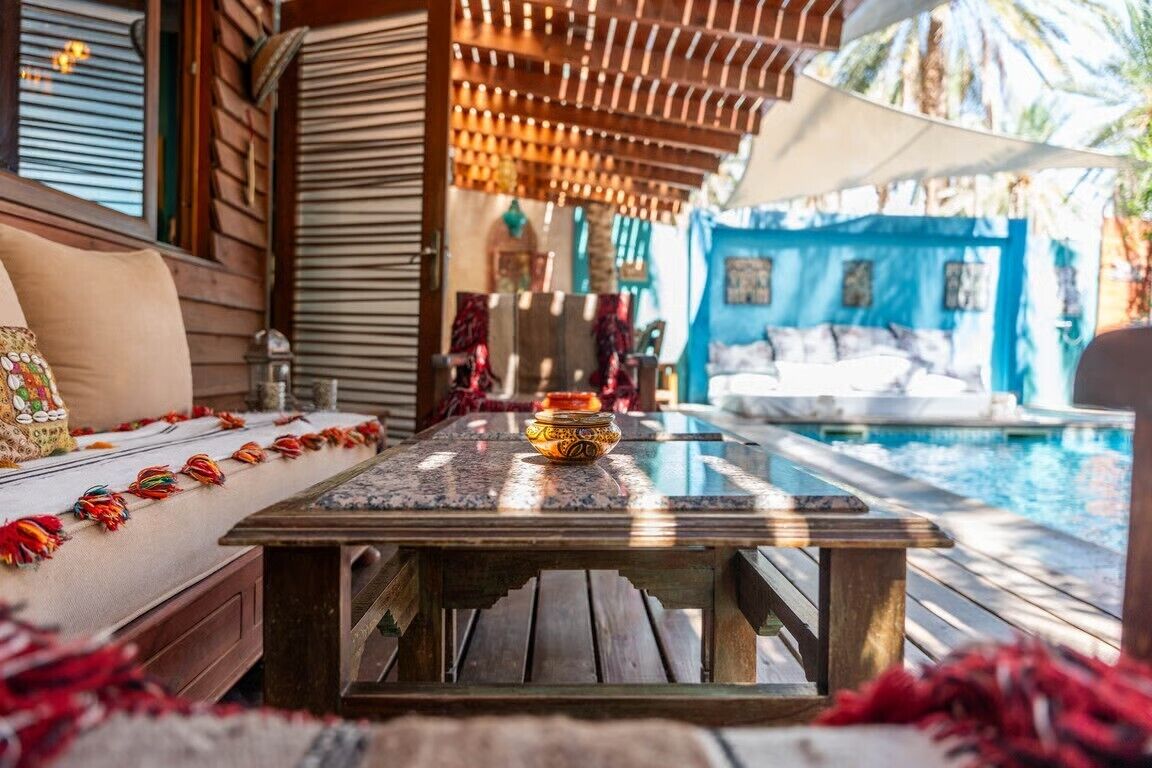 Una mesa de madera con un cuenco decorativo en el centro, un sofá acolchado con flecos coloridos y una piscina al fondo.