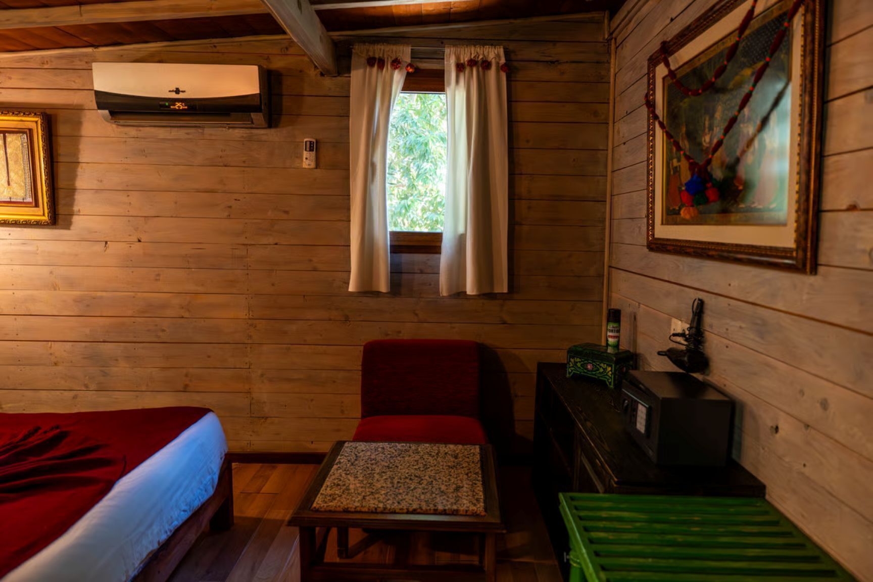 A cozy cabin room with a bed, a red chair, a small table, a green crate, a painting, and a window with curtains.