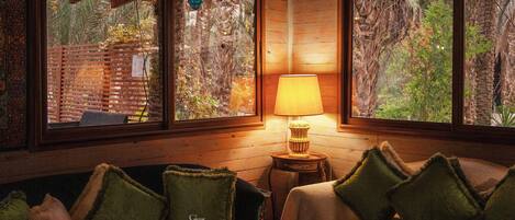 Una habitación acogedora con paredes de madera, un sofá con cojines verdes, una mesita con lámpara y ventanas amplias que ofrecen vistas a árboles y un muro de ladrillo.