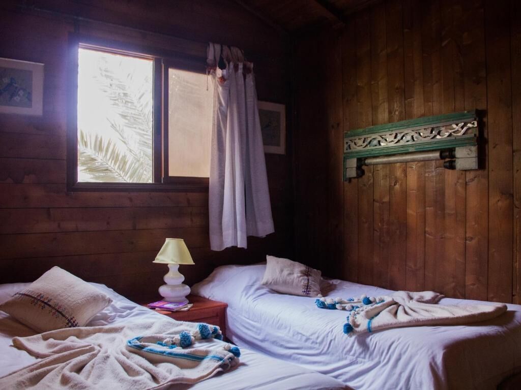 A cozy cabin room with two beds, a window with curtains, a lamp, and a wooden wall with a decorative bracket.