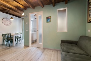 A room with a wooden ceiling, a dining table with chairs, a sofa, and a kitchen visible through an open door.