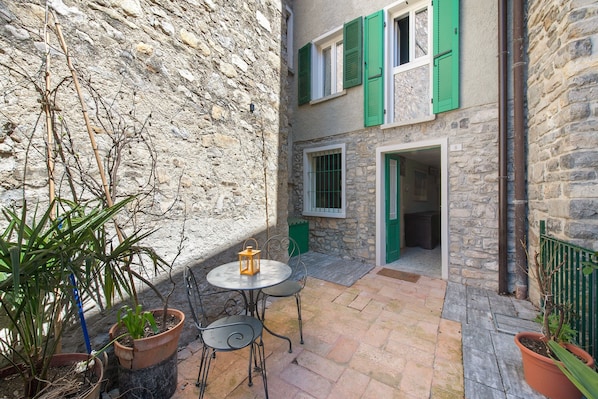 A stone building with green shutters, a small patio with a table and chairs, and potted plants.
