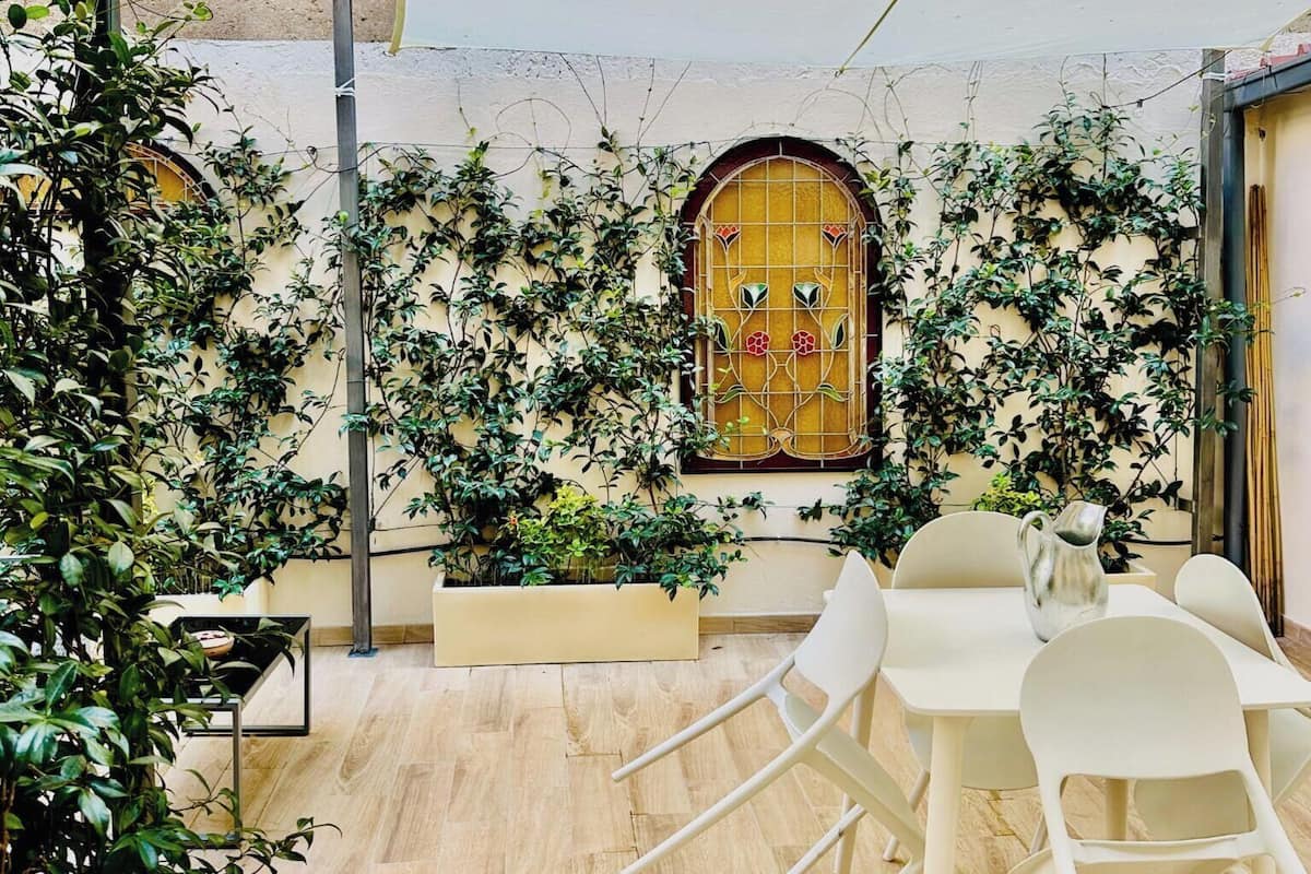 A courtyard with a white table and chairs, surrounded by lush greenery and a decorative window.