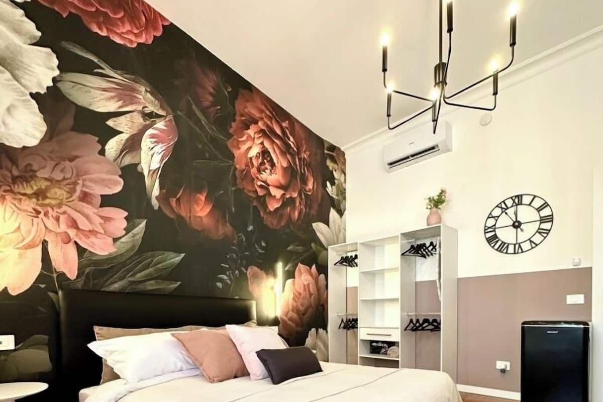 A bedroom with a large floral mural, a bed with white bedding, a black headboard, a bedside table, a wardrobe, a clock, and a small black appliance.