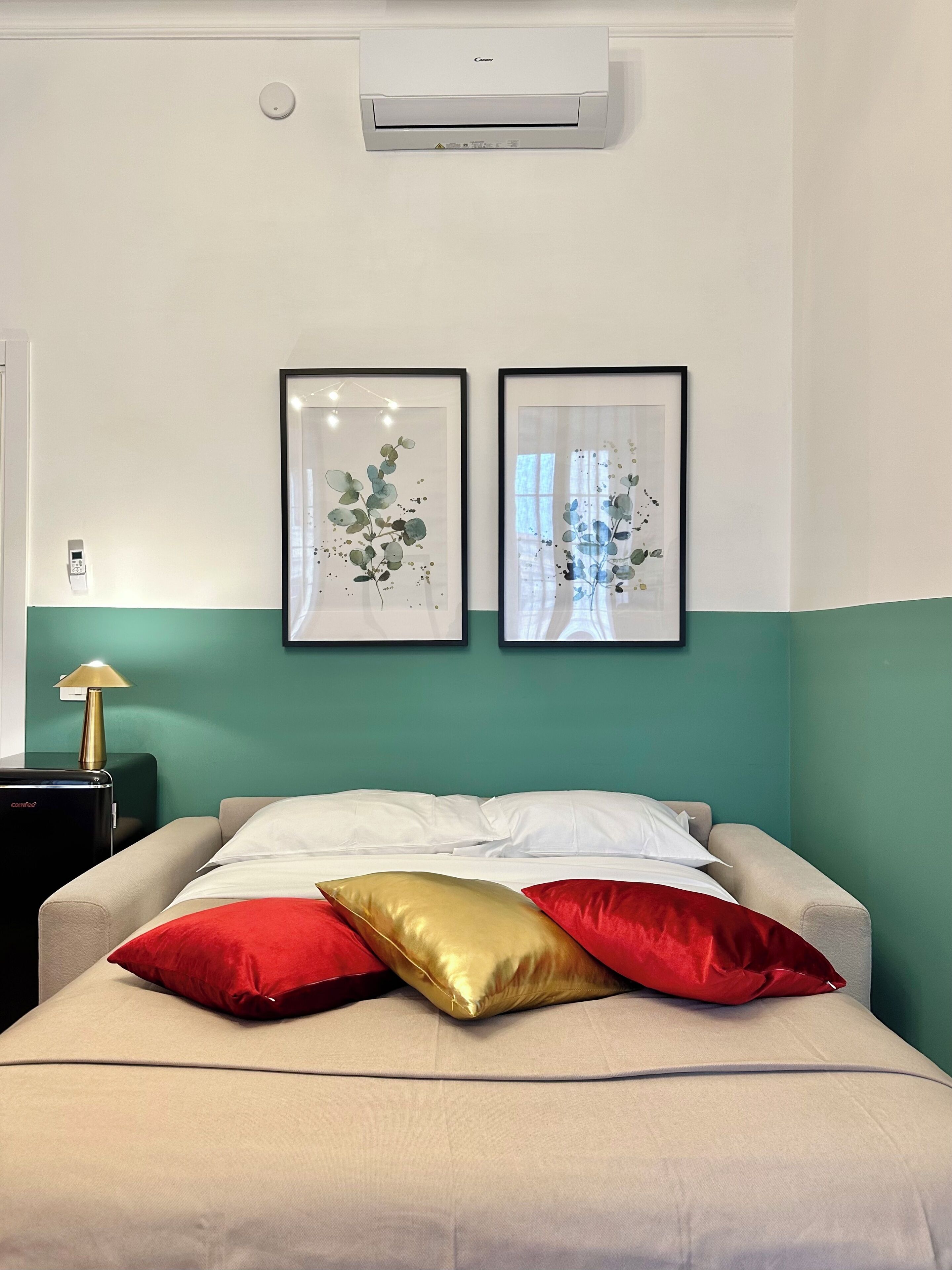 Une chambre d’hôtel avec un lit, deux reproductions botaniques encadrées accrochées au mur, une table de chevet avec une lampe et un appareil de climatisation placé au-dessus du lit.