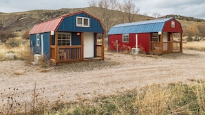 Two small cabins with metal roofs and wooden decks, set in a dry, grassy area with hills in the background.