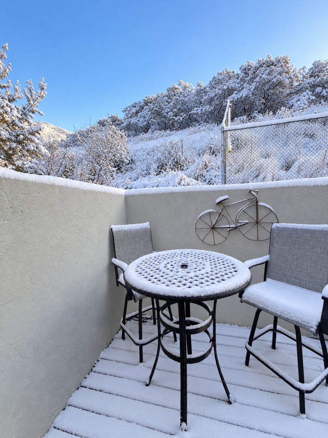 Eine verschneite Terrasse mit einem runden Tisch und zwei Stühlen, ein Fahrrad, das an der Wand lehnt, und ein Zaun, der das Areal umgrenzt.