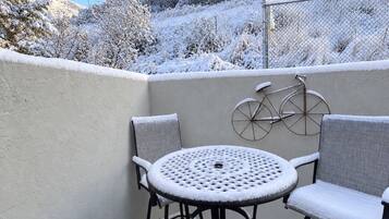 Eine verschneite Terrasse mit einem runden Tisch und zwei Stühlen, ein Fahrrad, das an der Wand lehnt, und ein Zaun, der das Areal umgrenzt.