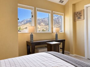 Un dormitorio con una cama, un escritorio con dos lámparas y vistas a montañas a través de tres ventanas.