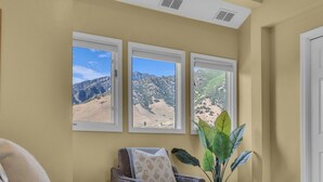 A bedroom with three windows offering views of a mountainous landscape, a bed with a patterned bedspread, a chair with a patterned pillow, and a large green plant. - Cottonwood Chalet - New Remodel - Private Hot Tub 3BR/2BA + 4 Ski Resorts (Cottonwood Heights)