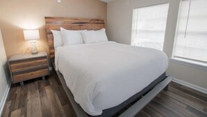 Un dormitorio con cabecera de madera, cubrecama blanco, mesita de noche con cajones y ventana con persianas.
