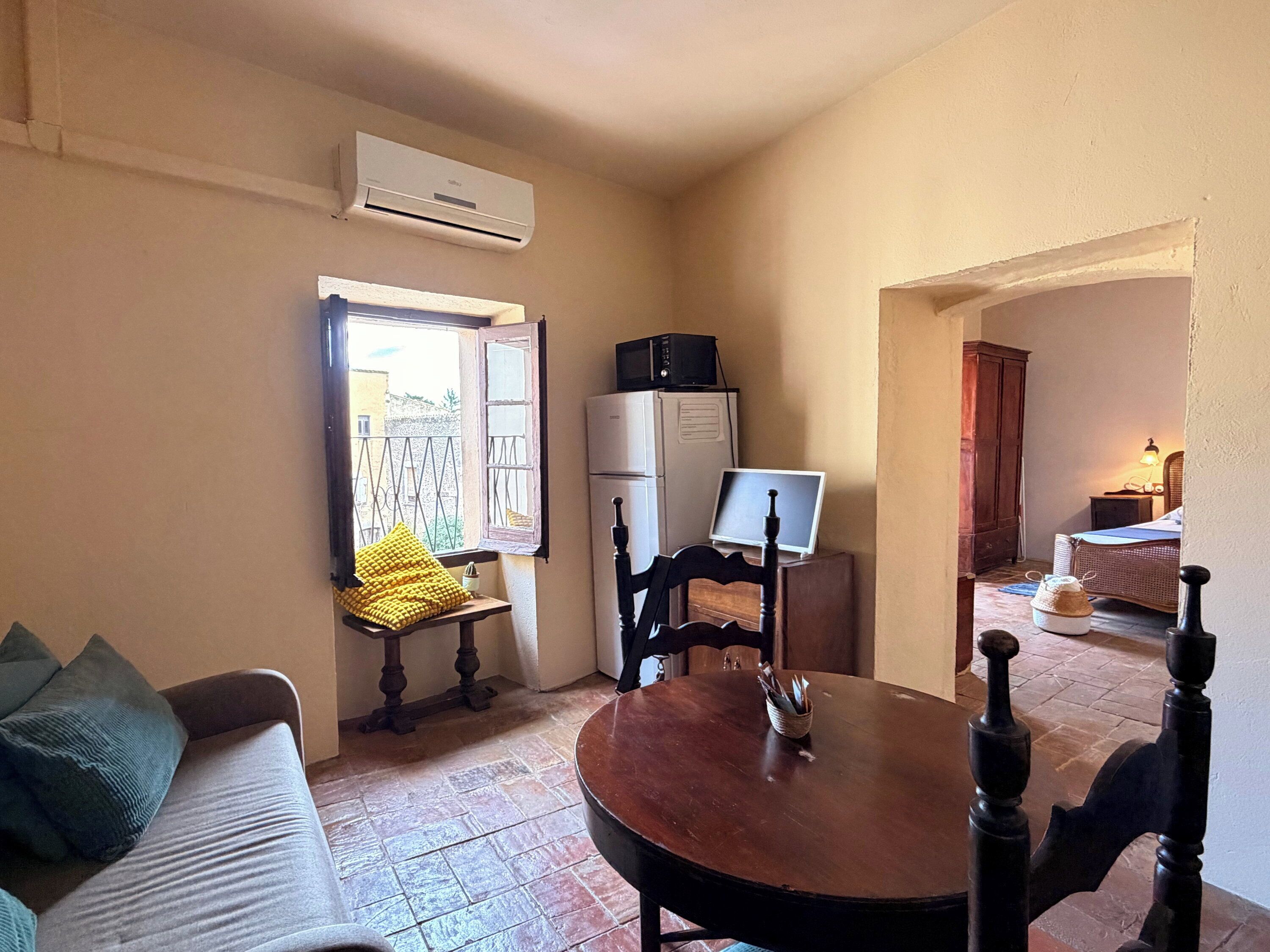 A cozy room with a wooden dining table, a sofa, a window with an open shutter, a white air conditioner, and a laptop on a cabinet.