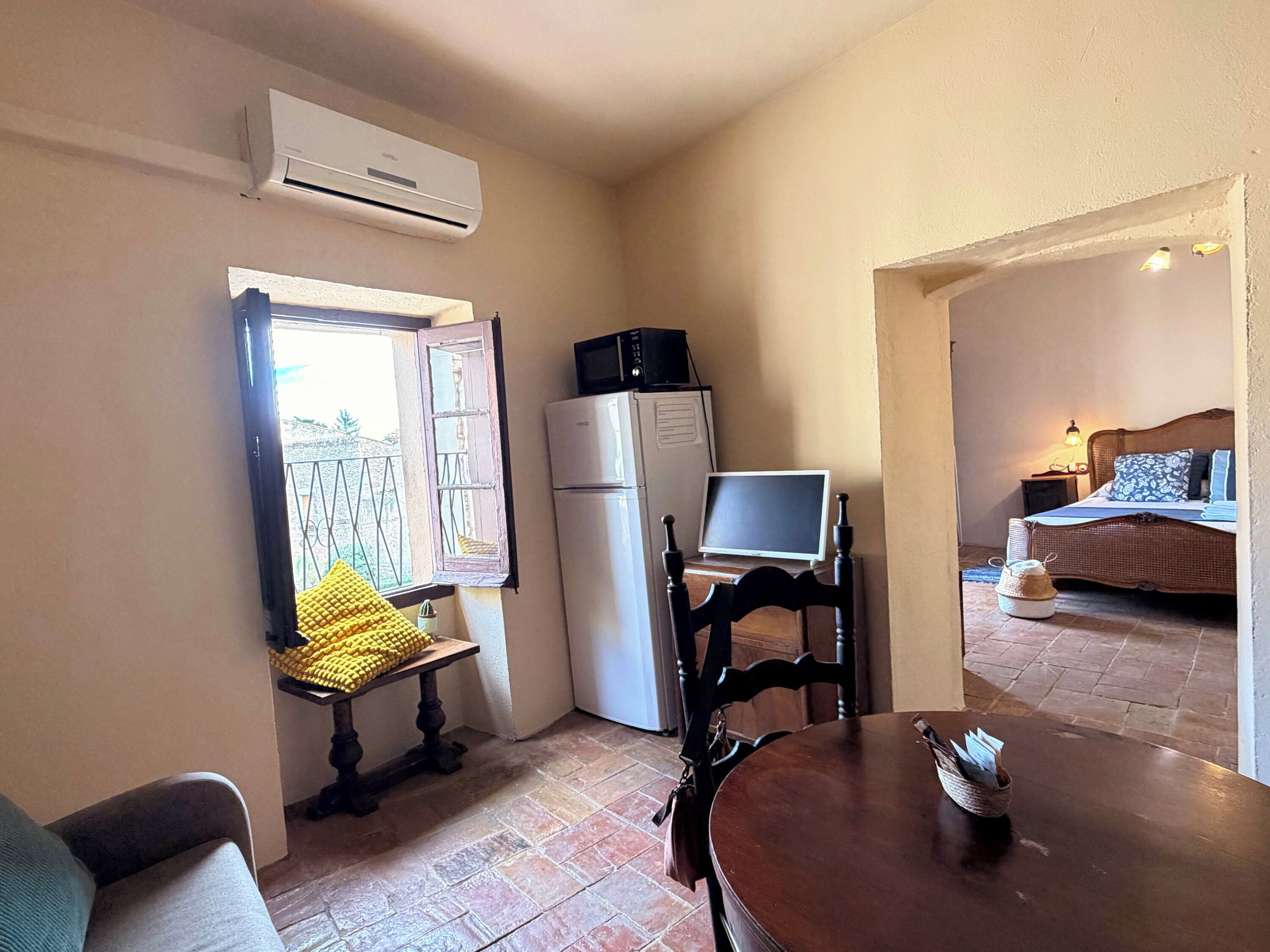 Ein kompaktes Zimmer mit Esstisch, einem kleinen Fenster mit Blick, einer Küchenzeile mit Kühlschrank und Mikrowelle sowie einem Bett im angrenzenden Raum.