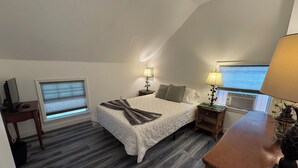 Une chambre à coucher comprenant un lit, deux tables de chevet avec des lampes, un bureau avec un ordinateur, une fenêtre avec des stores et un radiateur.
