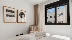 A modern hotel room with a bed, two framed artworks, a window with curtains, and a view of buildings outside. - Luxury Mirador Resort Apartment (Estepona)