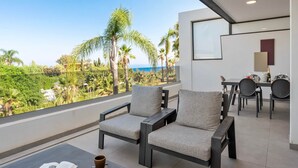 A balcony with two grey armchairs, a dining table with chairs, and a view of the ocean and greenery. - Luxury Mirador Resort Apartment (Estepona)