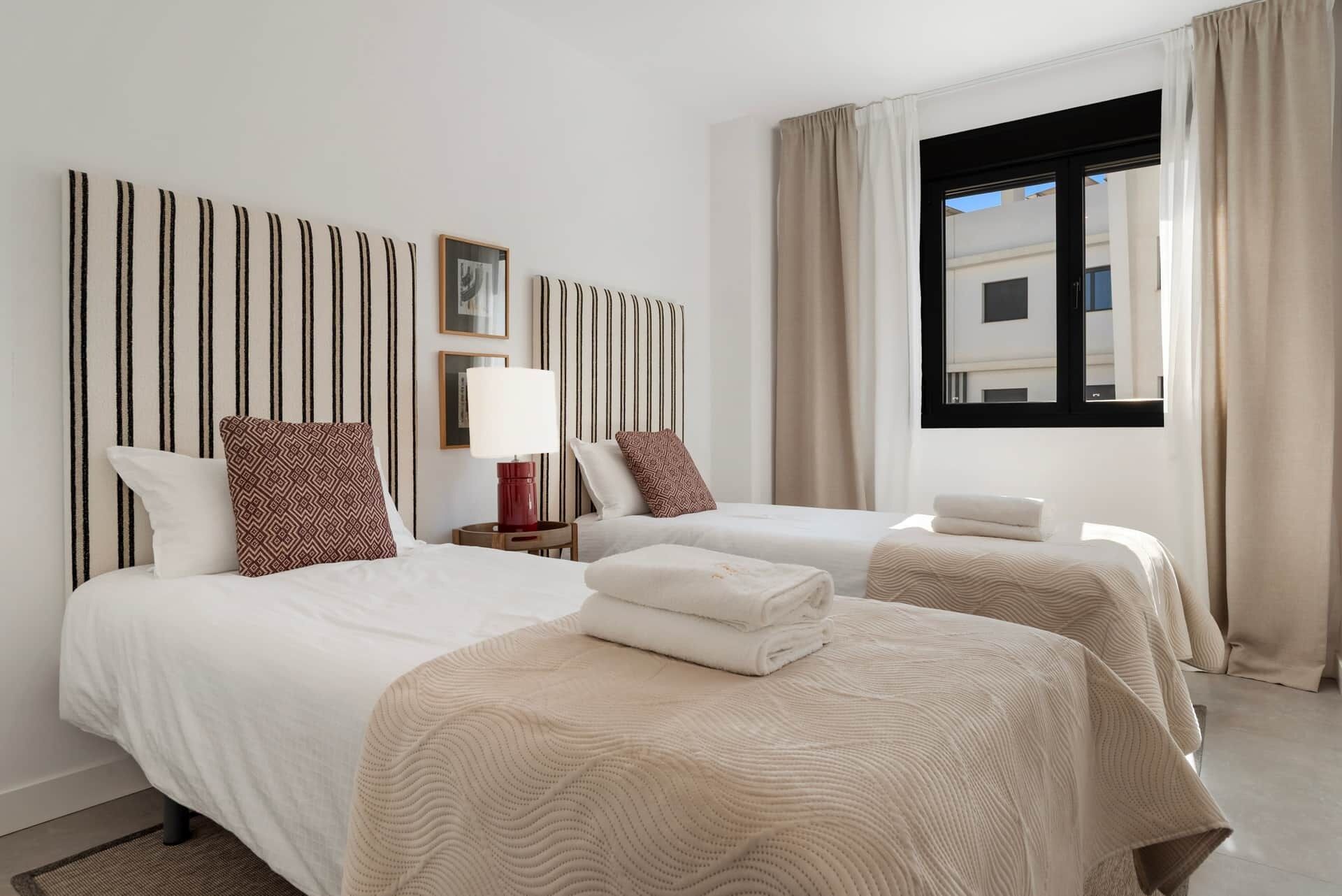 Habitació d'hotel amb dos llits, un panell del capçal estriat, una finestra amb cortines i una taula petita amb una làmpada.